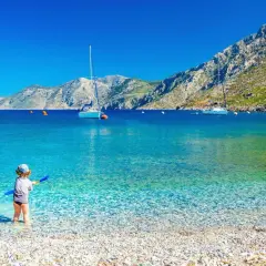 Dziecko bawi się na kamienistej plaży z turkusową wodą. W tle żaglówki i góry. Idealne miejsce, by znaleźć, gdzie najtaniej na wakacje.