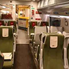 Wnętrze wagonu z zielonymi siedzeniami i stolikami. Widoczna numeracja miejsc w pociągu nad oknami.