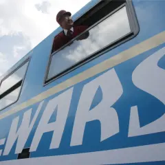 Konduktor w czerwonym mundurze wygląda z okna niebieskiego pociągu z napisem WARS.