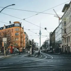 Skrzyżowanie w Berlinie z widoczną wieżą telewizyjną. Czy Berlin jest bezpieczny? Widać ludzi spacerujących, tramwaje i samochody.