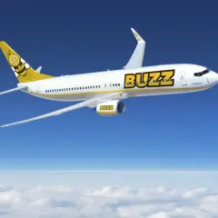 Samolot linii Buzz, należącej do Ryanair, z uśmiechniętą pszczołą na ogonie, leci na tle błękitnego nieba i chmur.