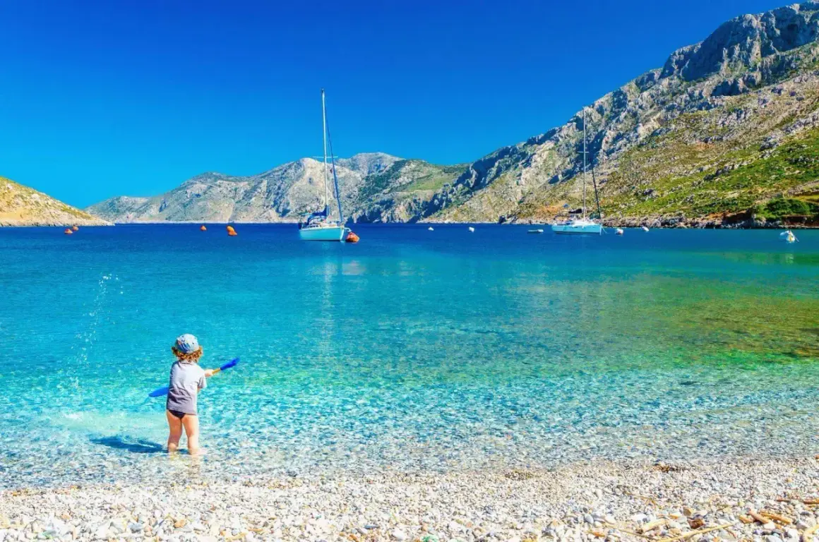 Dziecko bawi się na kamienistej plaży z turkusową wodą. W tle żaglówki i góry. Idealne miejsce, by znaleźć, gdzie najtaniej na wakacje.