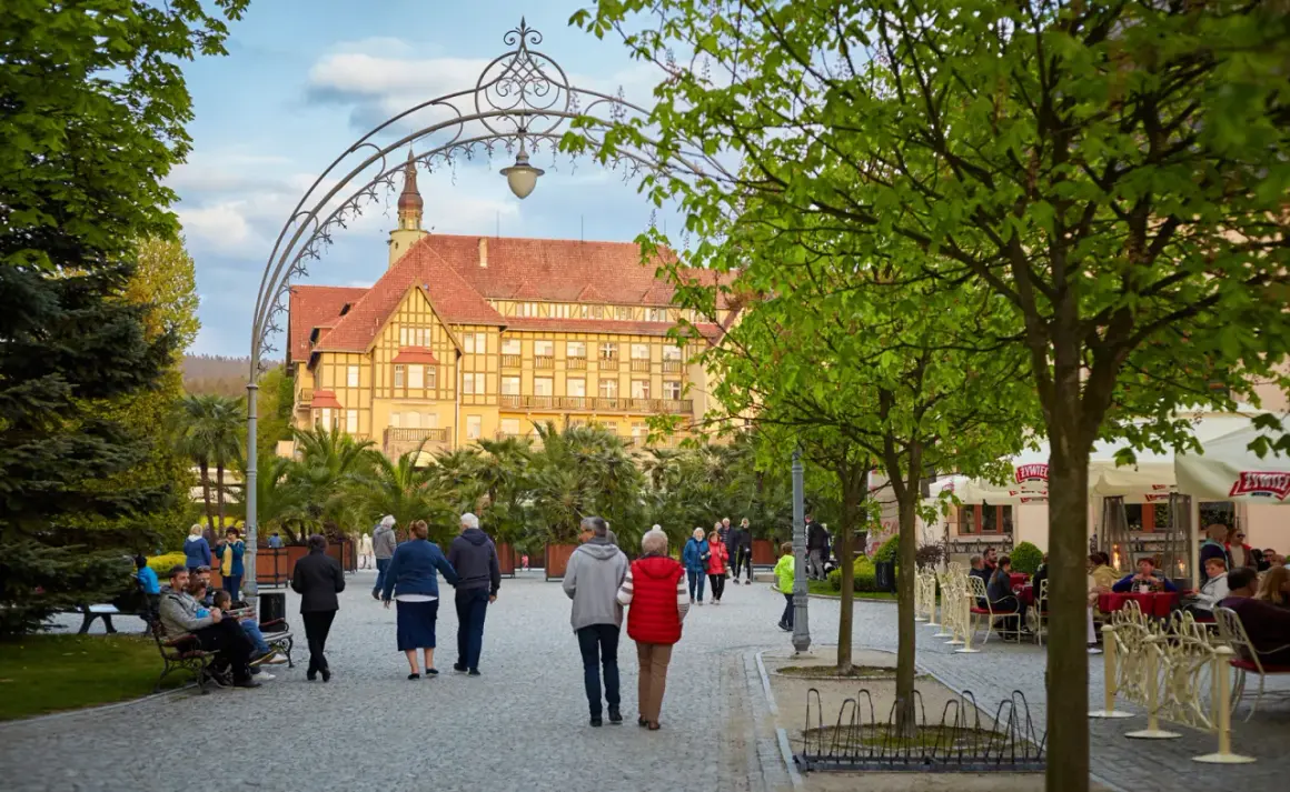 Wieczorny spacer po Kudowie-Zdroju. Ludzie przechadzają się alejką, podziwiając architekturę i zielone otoczenie.