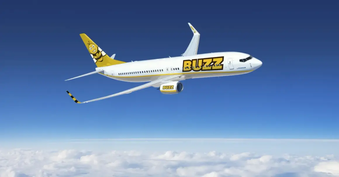 Samolot linii Buzz, należącej do Ryanair, z uśmiechniętą pszczołą na ogonie, leci na tle błękitnego nieba i chmur.