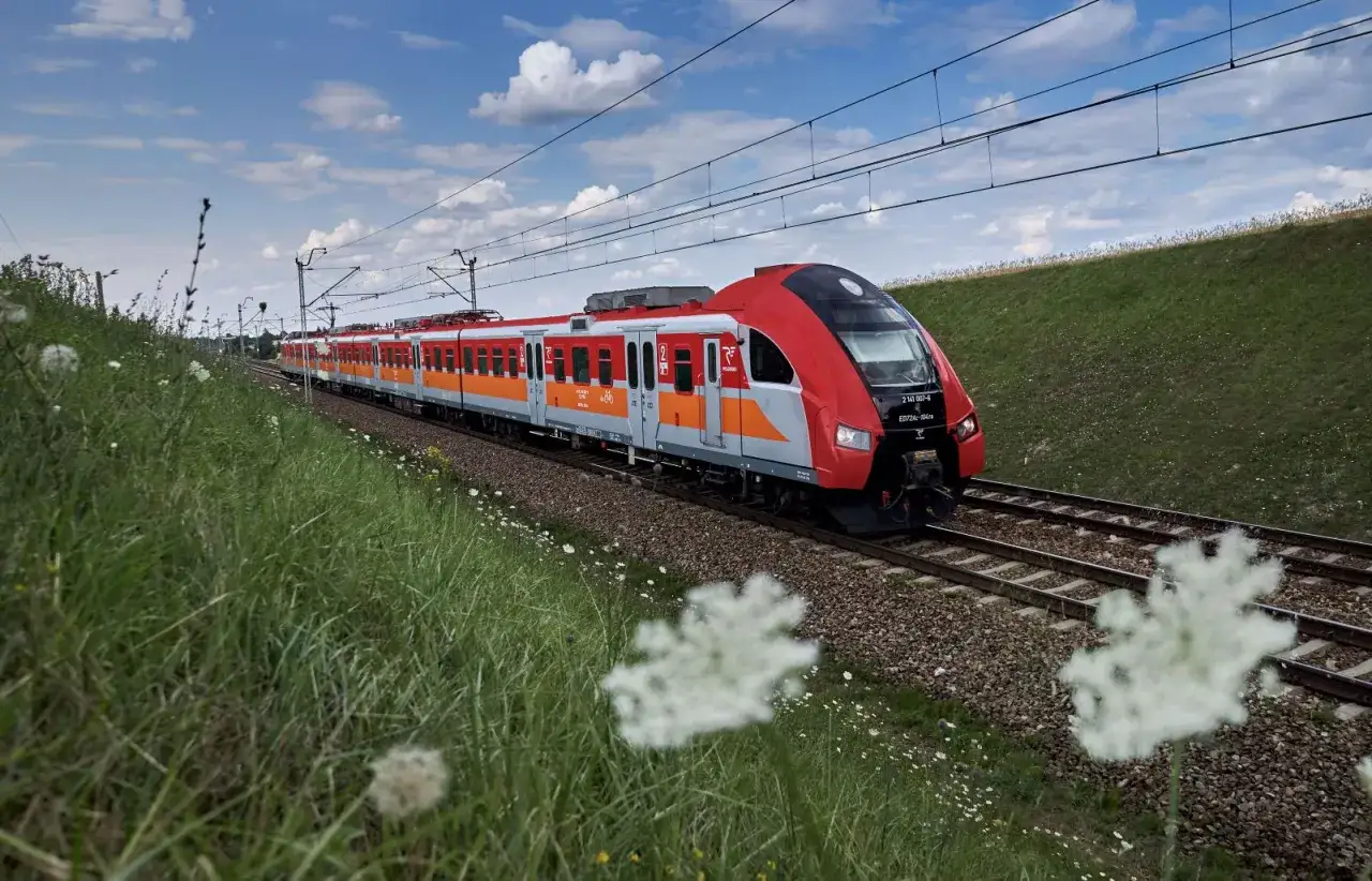 Czerwono-pomarańczowy pociąg Polregio jedzie po torach wśród zieleni. Idealny na wycieczkę z biletem turystycznym Polregio.