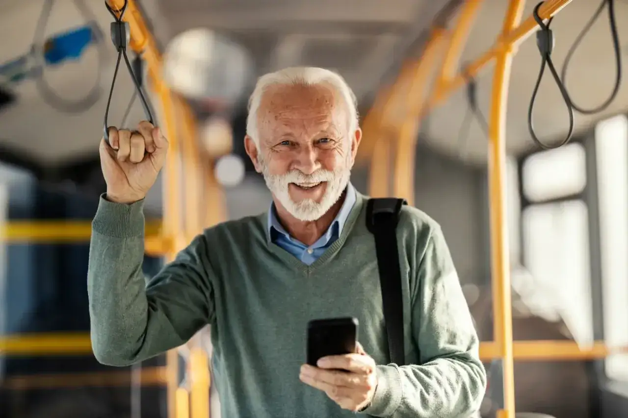 Uśmiechnięty emeryt trzymający się poręczy w autobusie, z telefonem w dłoni. Czeka na swój przejazd PKP, mając przy sobie zaświadczenie dla emeryta na przejazd PKP.