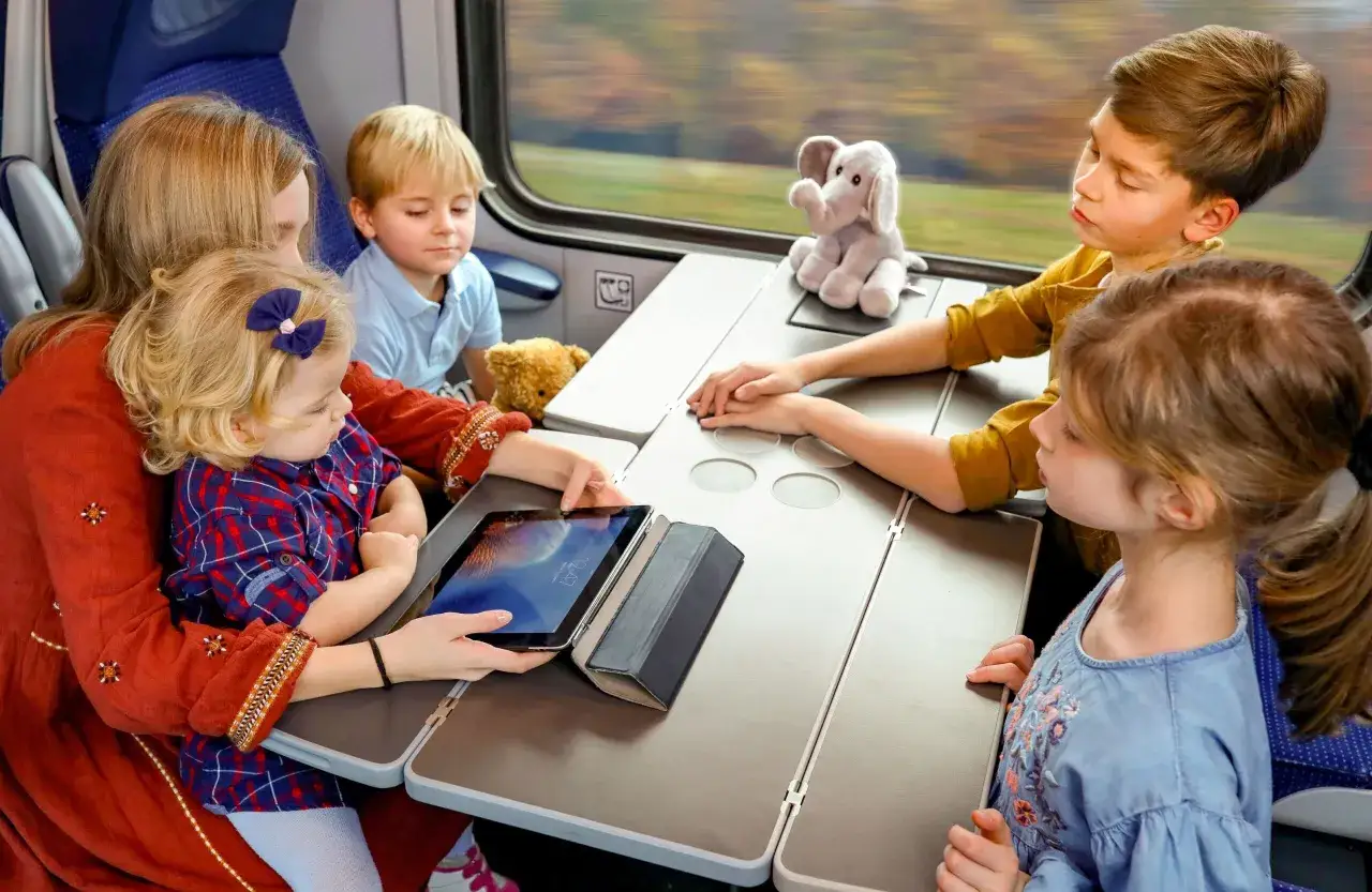 Rodzina z dziećmi podróżuje pociągiem PKP. Dzieci oglądają bajki na tablecie, a mama trzyma go w rękach.