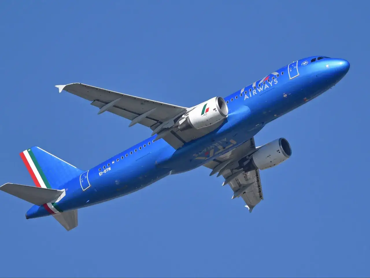 Niebieski samolot ITA Airways z włoską flagą na ogonie wznosi się na tle błękitnego nieba.