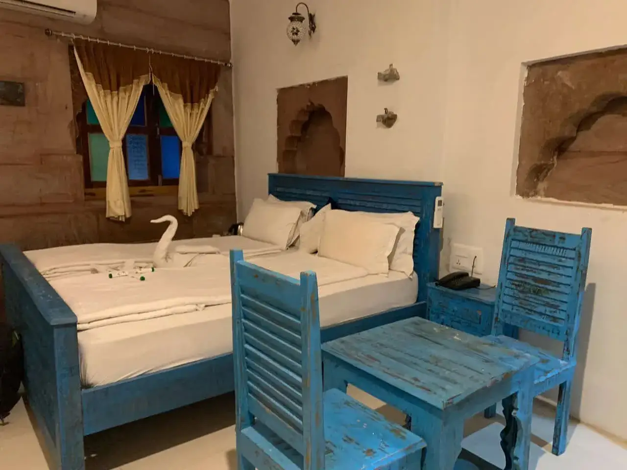 Pokój hotelowy w Indiach z łóżkiem i stolikiem w kolorze niebieskim. Uroczy łabędź z ręcznika na pościeli. Ceny w Indiach mogą być zaskakująco przystępne.
