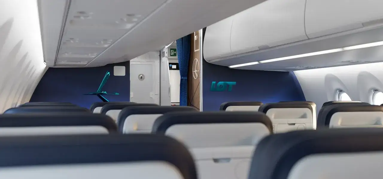 Wnętrze samolotu Airbus A220 linii LOT. Widok na rzędy siedzeń i logo LOT na granatowej ścianie.
