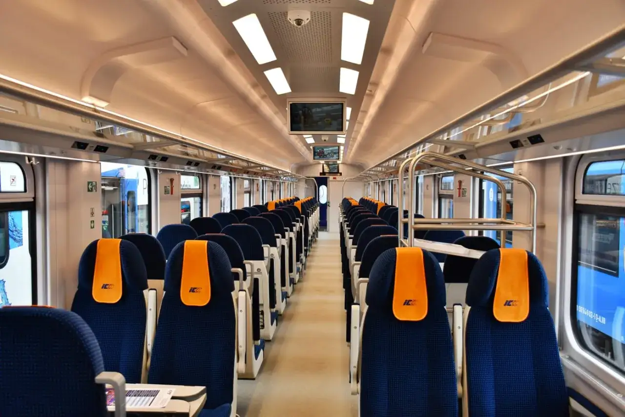 Wnętrze nowoczesnego pociągu PKP Intercity, z rzędami niebieskich foteli z pomarańczowymi zagłówkami, gotowego do podróży do Zamościa.