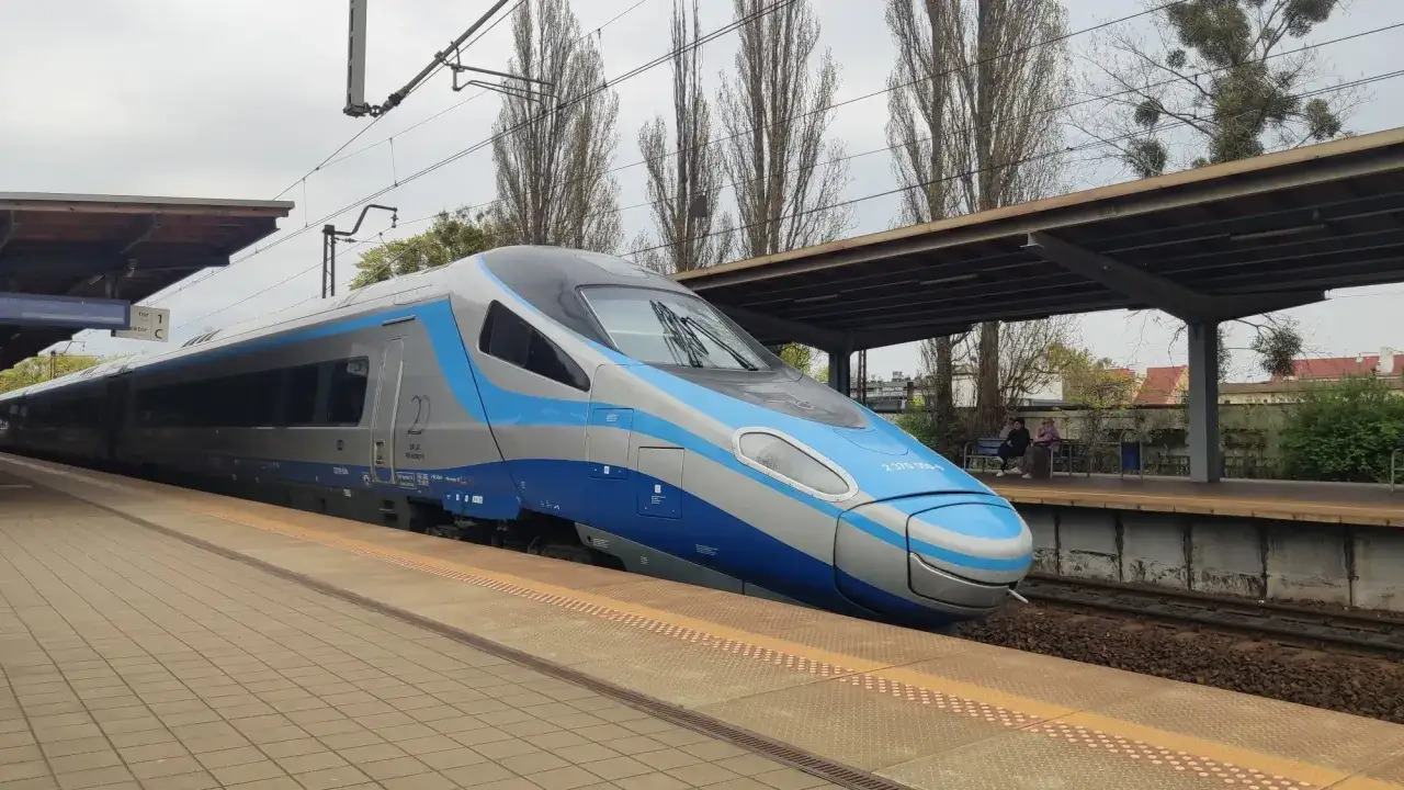 Nowoczesny pociąg Pendolino z niebieskimi i szarymi pasami stoi na peronie. W tle widać drzewa i budynki. To idealny środek transportu, by szybko i komfortowo podróżować.