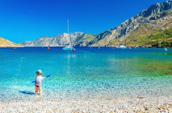 Dziecko bawi się na kamienistej plaży z turkusową wodą. W tle żaglówki i góry. Idealne miejsce, by znaleźć, gdzie najtaniej na wakacje.