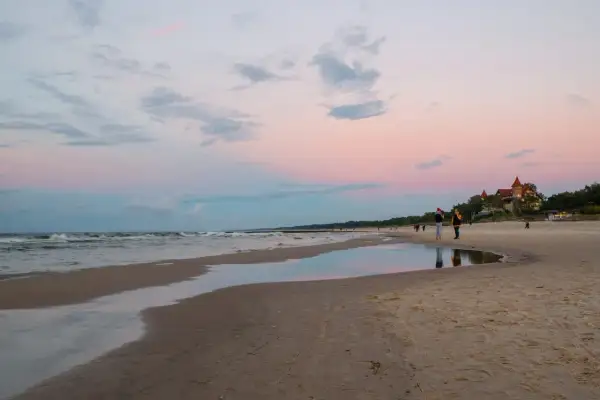 Zachód słońca nad morzem w Łebie. Kilka osób spaceruje po plaży, a w oddali widać budynek przypominający zamek.