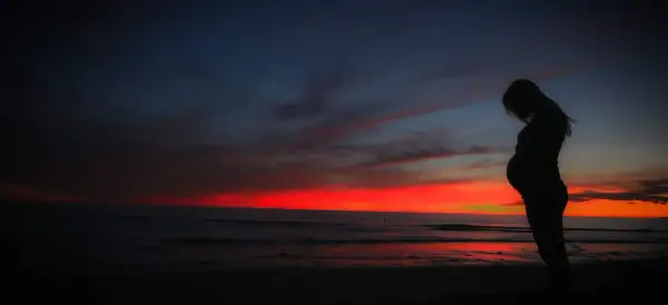 Kobieta w ciąży na tle zachodu słońca nad oceanem. Pamiętaj o ubezpieczeniu turystycznym w ciąży, by podróżować bezpiecznie.