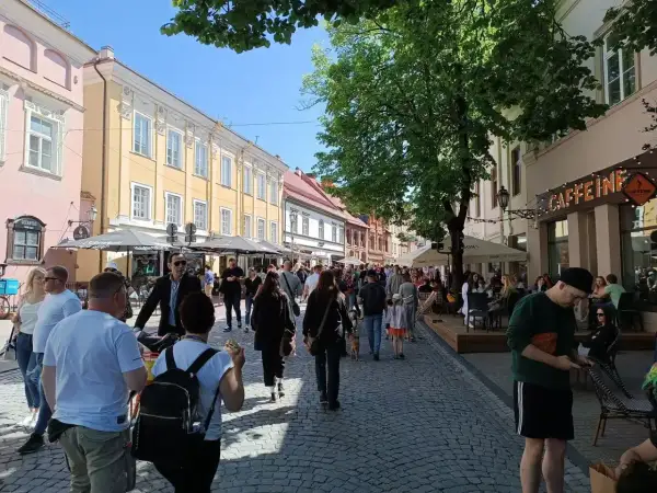 Tłum ludzi spaceruje po brukowanej ulicy w Wilnie, podziwiając architekturę i kawiarnie. Wygląda na to, że czy na Litwie jest bezpiecznie, bo atmosfera jest swobodna i radosna.