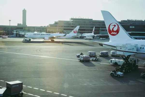 Samoloty Japan Airlines na lotnisku w Japonii. W tle budynki terminali i wieża kontroli lotów. Na płycie lotniska widać obsługę naziemną i ładunki.